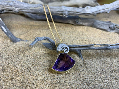 Gold pendant with a fan-shaped purple stone under a spiral, on driftwood