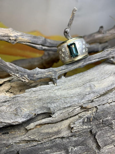 Wide ring with an emerald-cut green-blue stone and small diamonds, on driftwood