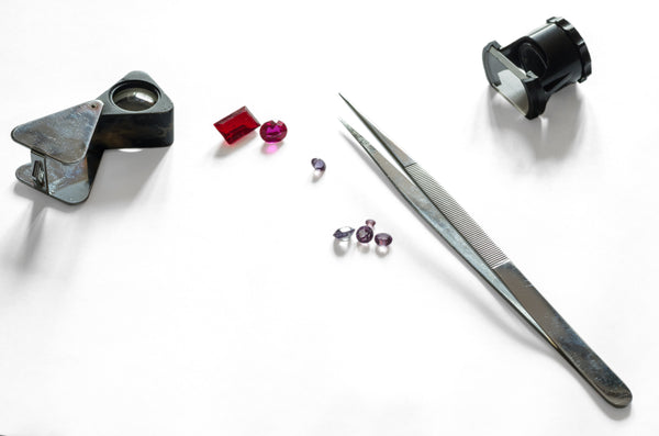A loupe, tweezers and loose pink and red gemstones on a white surface