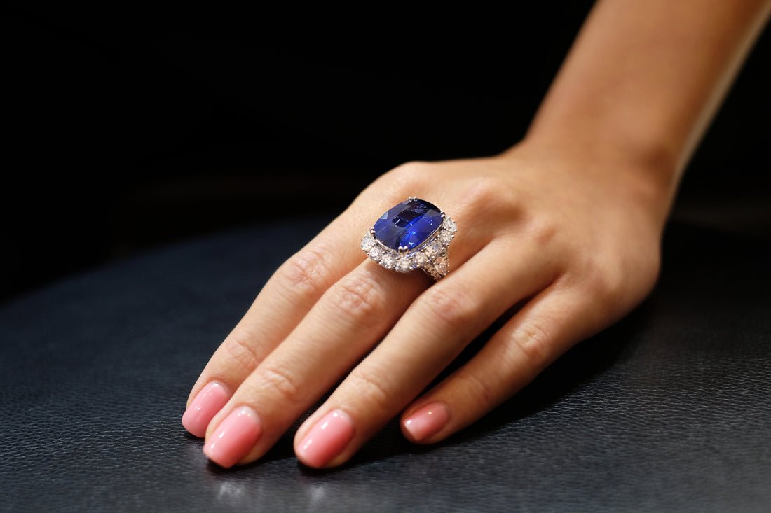 A hand wearing a ring with an oval blue stone in a halo of small diamonds